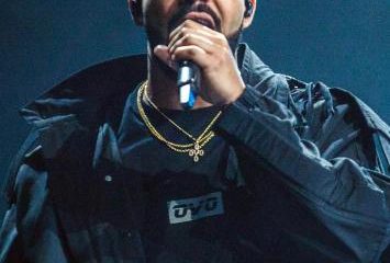 Drake performing on stage in a dark jacket, holding a microphone